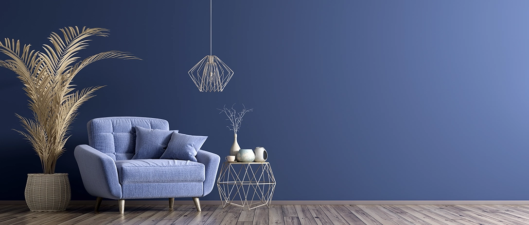 A living room with cozy blue armchair, decorative pillows, a geometric side table, and a plant in front of a deep blue wall, with wooden flooring adding warmth to the space.