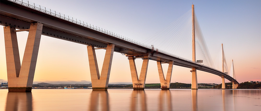 A modern cable-stayed bridge with tall, V-shaped concrete supports stretches across a calm river at sunset, reflecting softly on the water's surface.