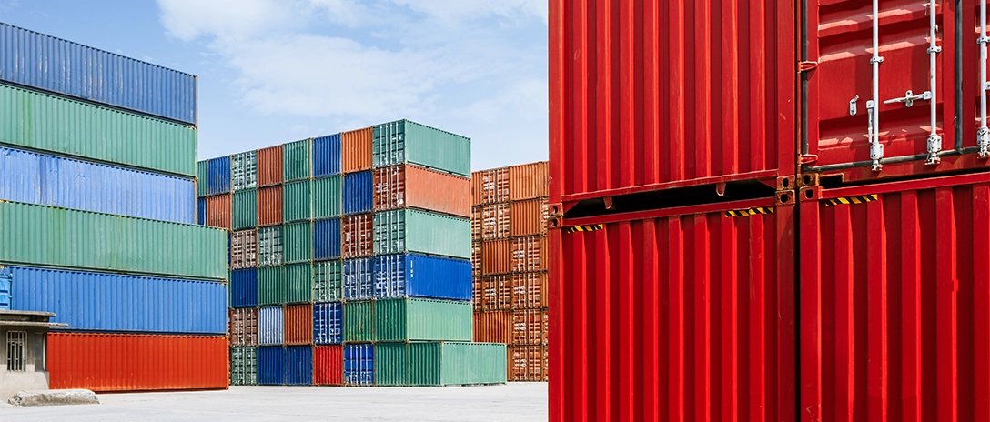 Stacks of brightly colored shipping containers, primarily in red, blue, green, and orange, are neatly arranged in a large open area under a clear sky.