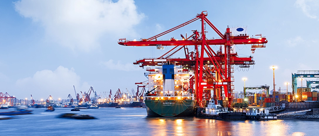 A large cargo ship is being loaded by towering red cranes at a busy port, with lights reflecting off the water under a partly cloudy evening sky.