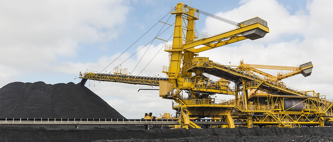A large yellow mining machine is actively loading coal onto a massive pile at a mining site, under a cloudy sky.