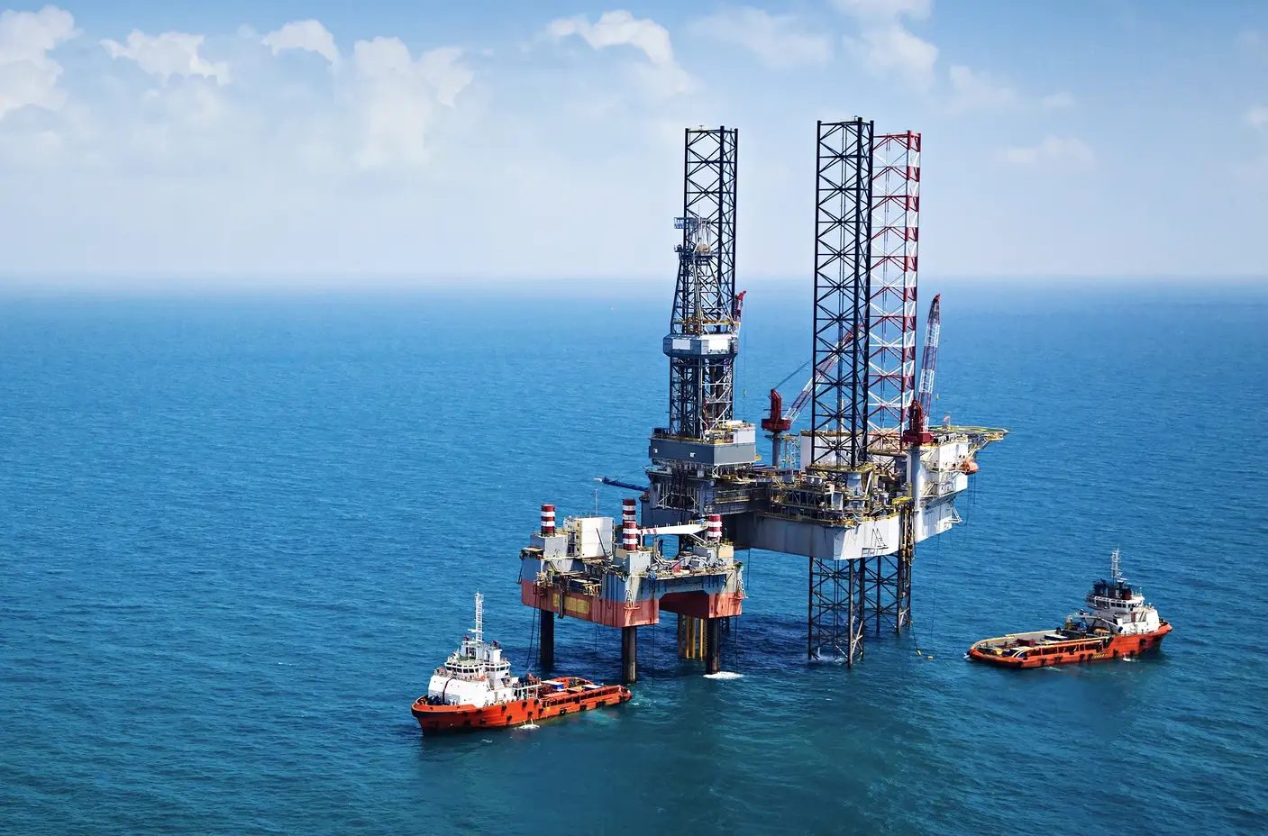An offshore oil platform stands in the middle of the ocean, supported by two tugboats, with calm blue waters stretching to the horizon under a partly cloudy sky.