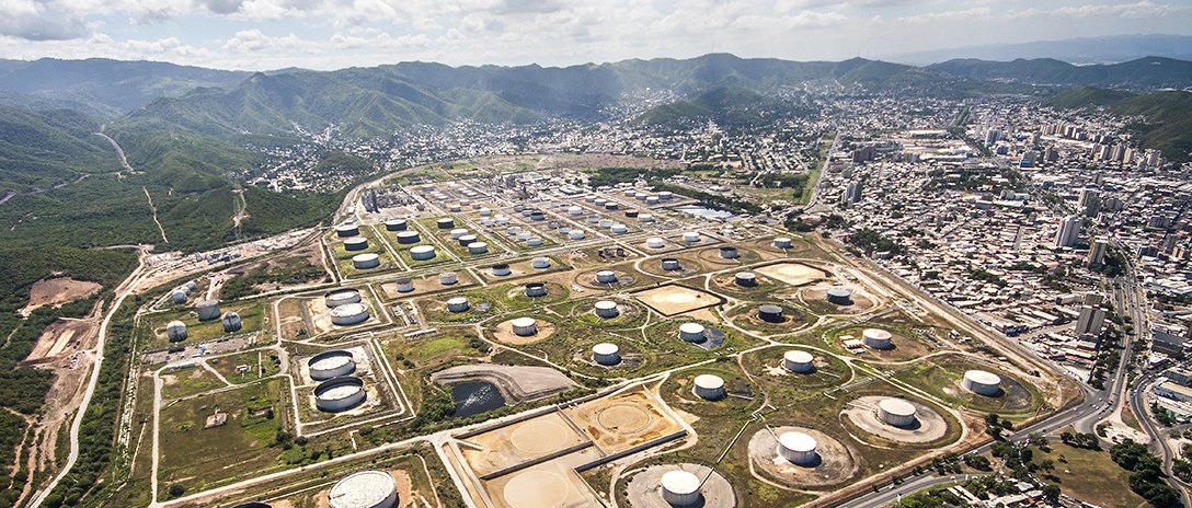 A large oil storage facility with numerous storage tanks is spread out across a large area, surrounded by mountains and a nearby city under a partly cloudy sky.