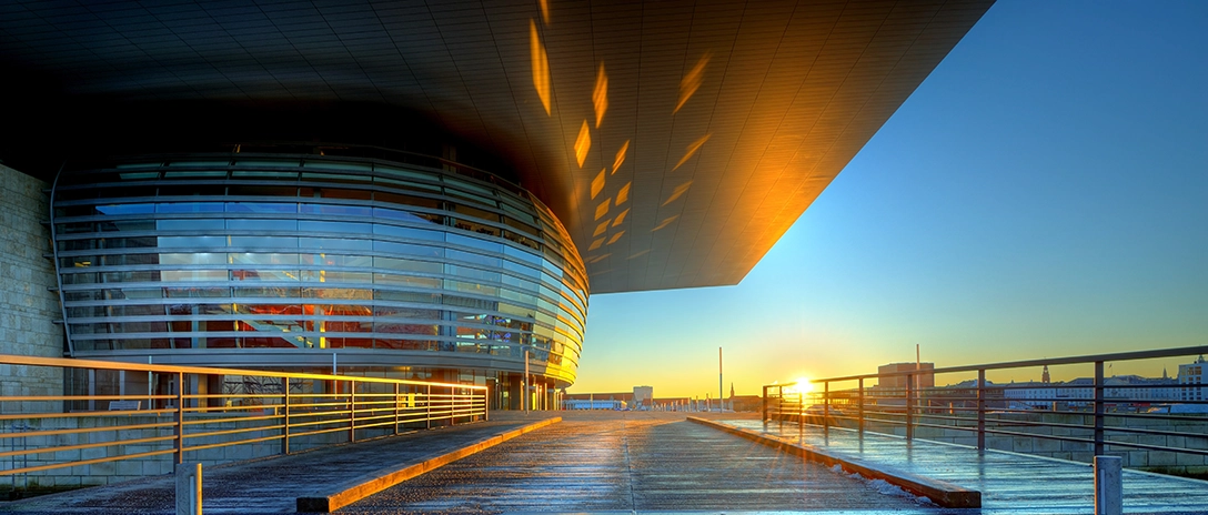 The Oslo Opera House showcases a curved glass facade and overhanging roof, glowing in the light of the sunset.