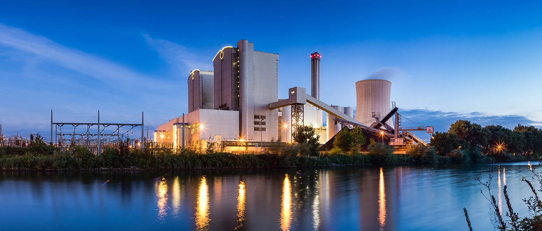 A power plant, illuminated at dusk, sits by a calm river with its lights reflecting on the water's surface.