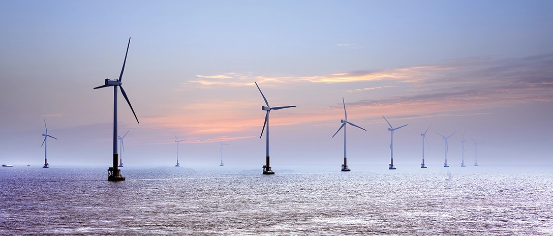 A row of offshore wind turbines stands in the calm sea at dusk, with the soft colors of the sunset fading into the horizon.
