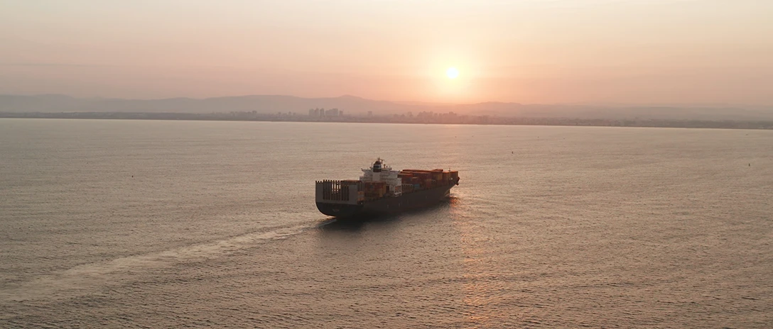 A container ship sailing on calm water toward the horizon during a sunset