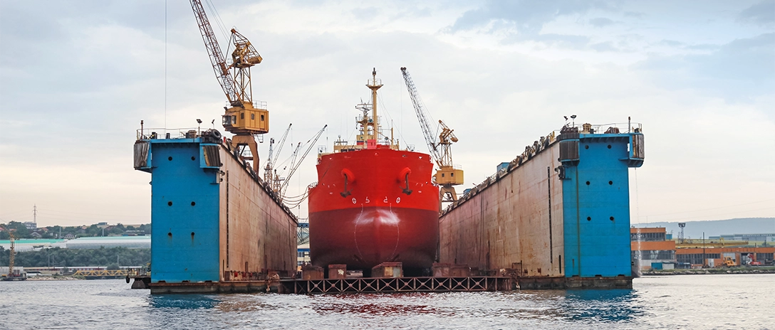 A large red ship sits in a dry dock between two massive blue walls, surrounded by cranes and industrial equipment, as it undergoes maintenance or repairs.