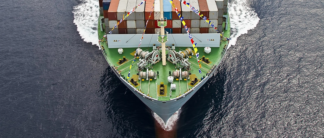 A large container ship, decorated with colorful flags, sails through the ocean, viewed from above with its bow cutting through the water.