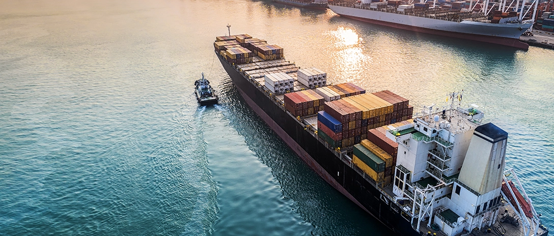 A large container ship, loaded with multicolored shipping containers sails through calm waters near a port at sunset.