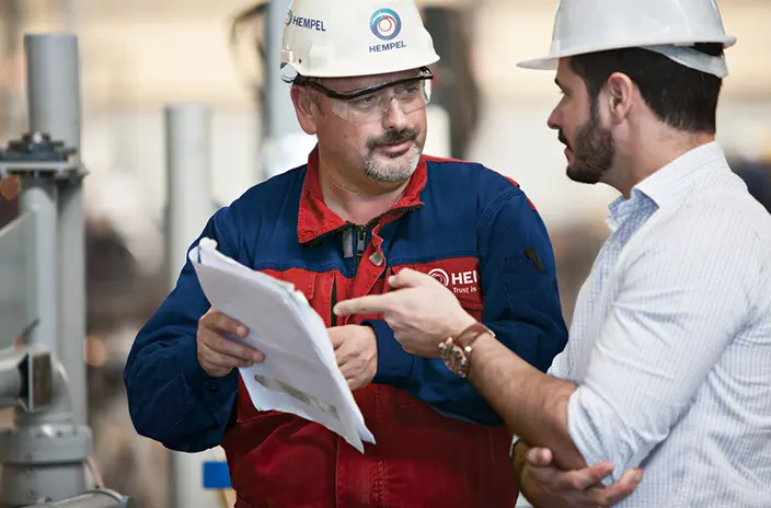 A Hempel coating advisor in a protective suit and helmet discusses paperwork with a colleague on a worksite.