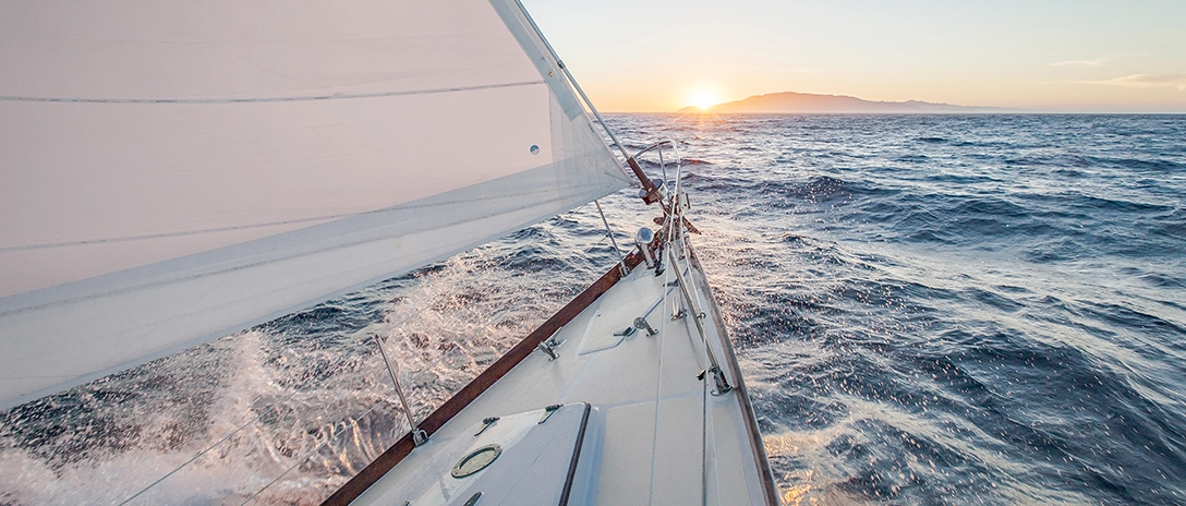 A yacht cuts through the waves at sunset, its sails full of wind, with an island in the distance.