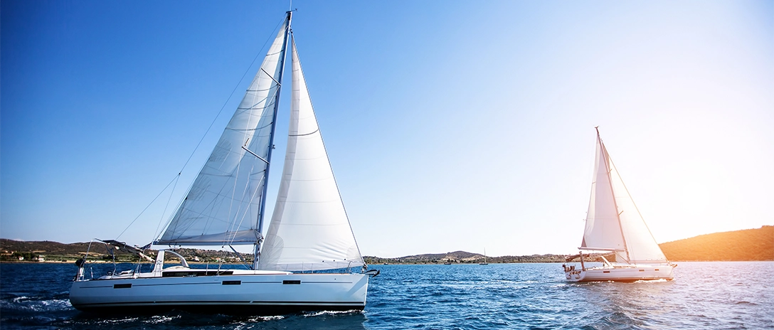 Two white yachts sail on calm blue waters under a clear sky, with low hills visible in the background