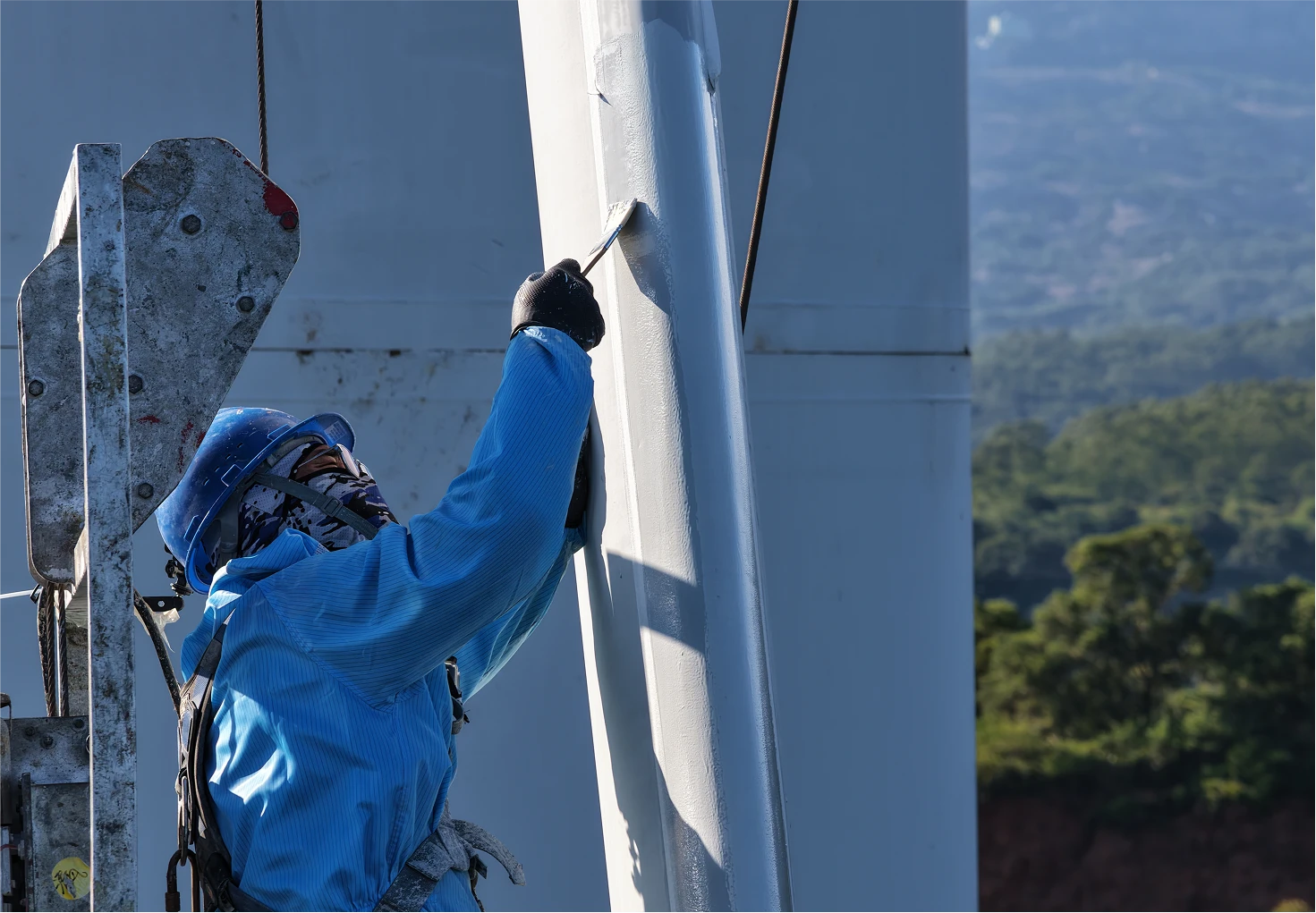 Worker wearing safety gear applies coating on a wind turbine blade while suspended on a platform.
