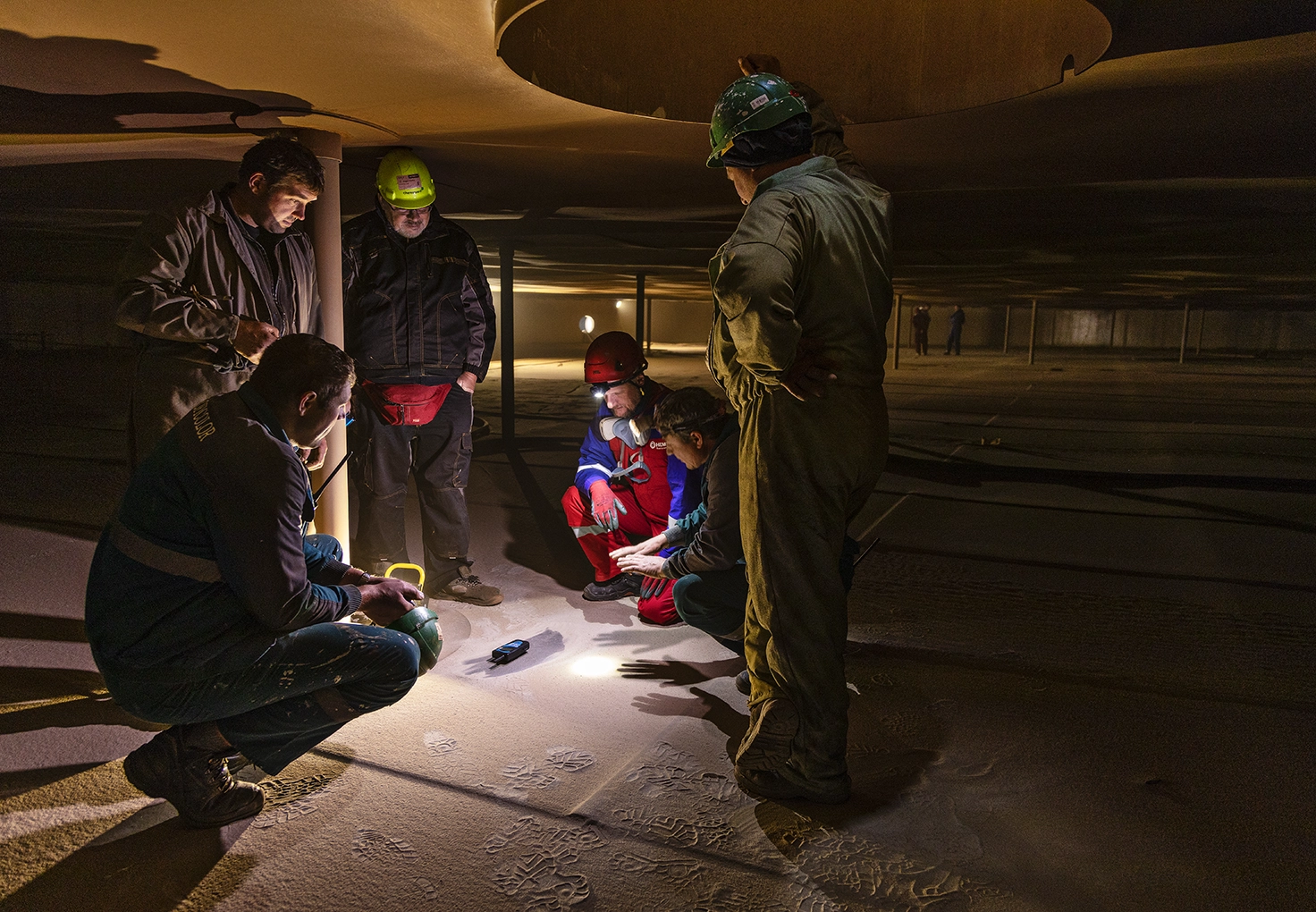 Workers gather inside an oil tank, analyzing a sample illuminated by a flashlight.