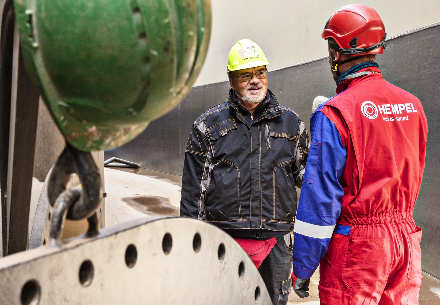 Two workers in protective gear discuss the coating process in an industrial setting.