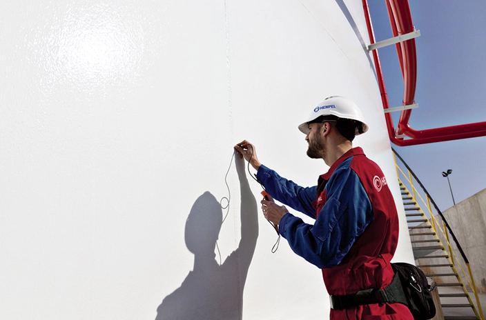 A Hempel inspector checks the integrity of a white-coated tank with a tool.
