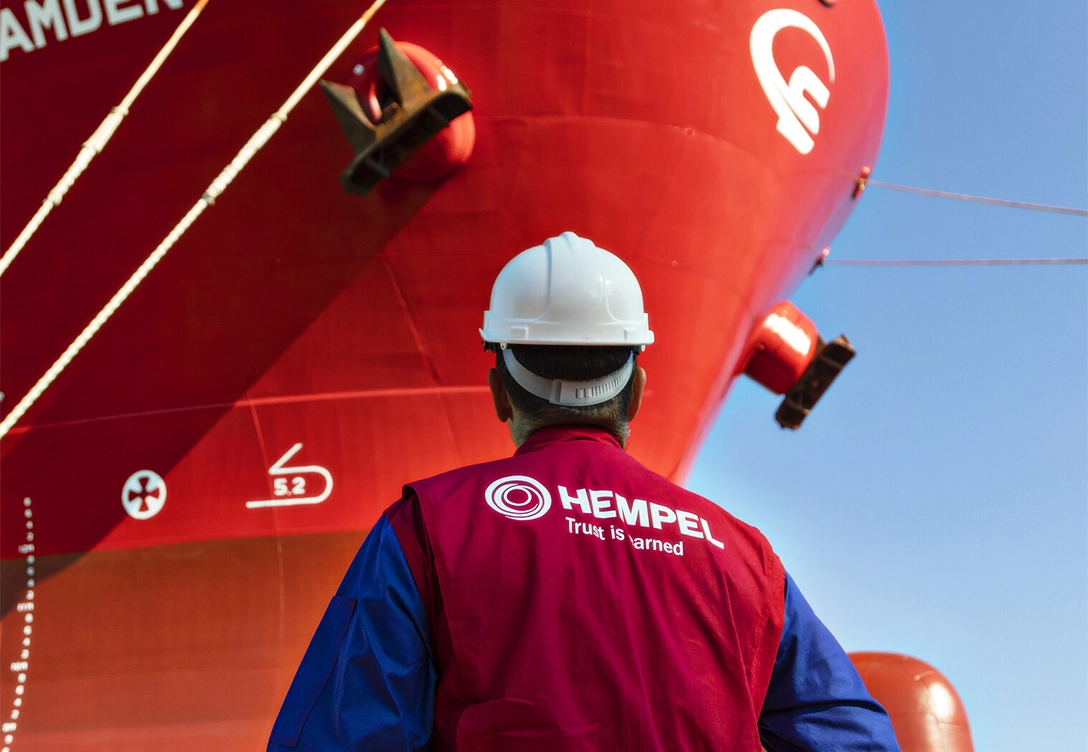 A Hempel worker inspects the exterior of a large red ship at a dock, wearing safety gear and a Hempel vest.