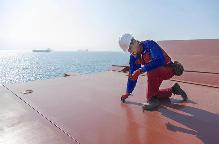 A Hempel worker kneeling on the deck of a ship at sea, using a tool to measure the coating thickness of the ship’s surface, with the ocean in the background.