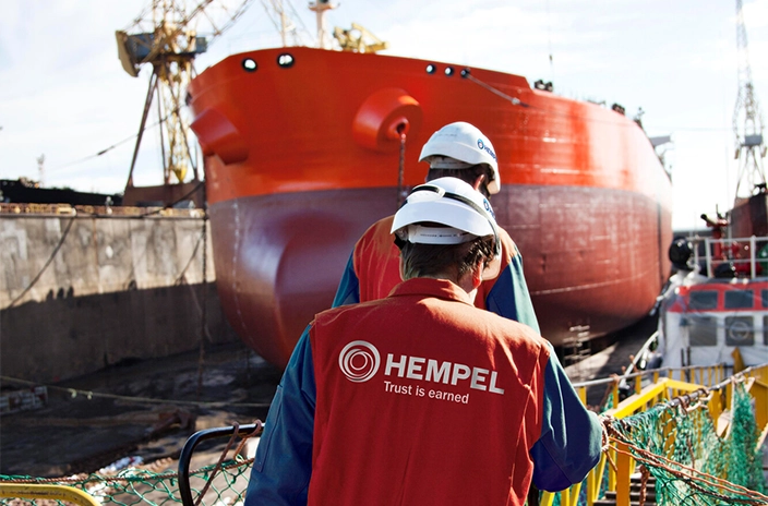 Two Hempel coating advisors in red uniforms and helmets work near a large red ship in a dock.