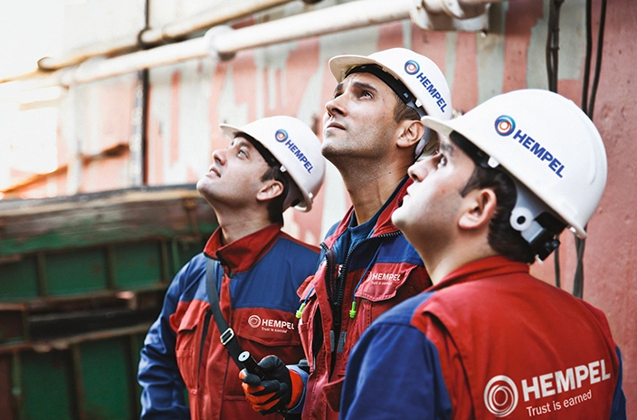 Three Hempel advisors in helmets and red uniforms look upward, inspecting the area around them.