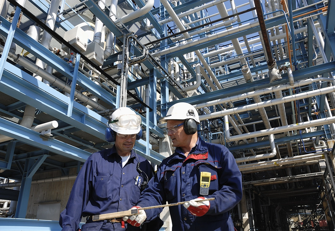 Two workers in safety gear review a wooden scale for measuring while standing in an industrial facility with blue pipe structures.