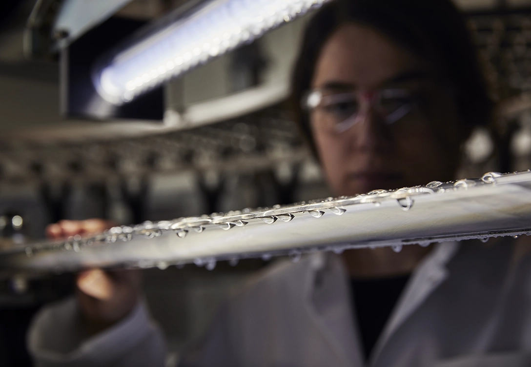 A woman in safety glasses inspecting a metal surface with water droplets during a rain erosion test in a laboratory.