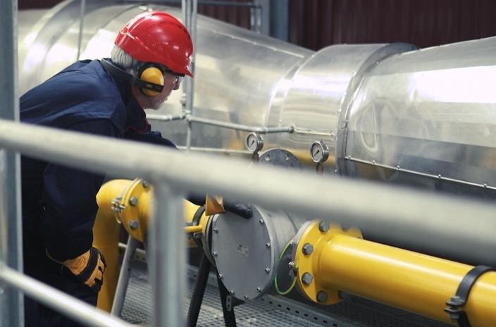 A worker wearing a red helmet and protective gear inspecting large pipes.