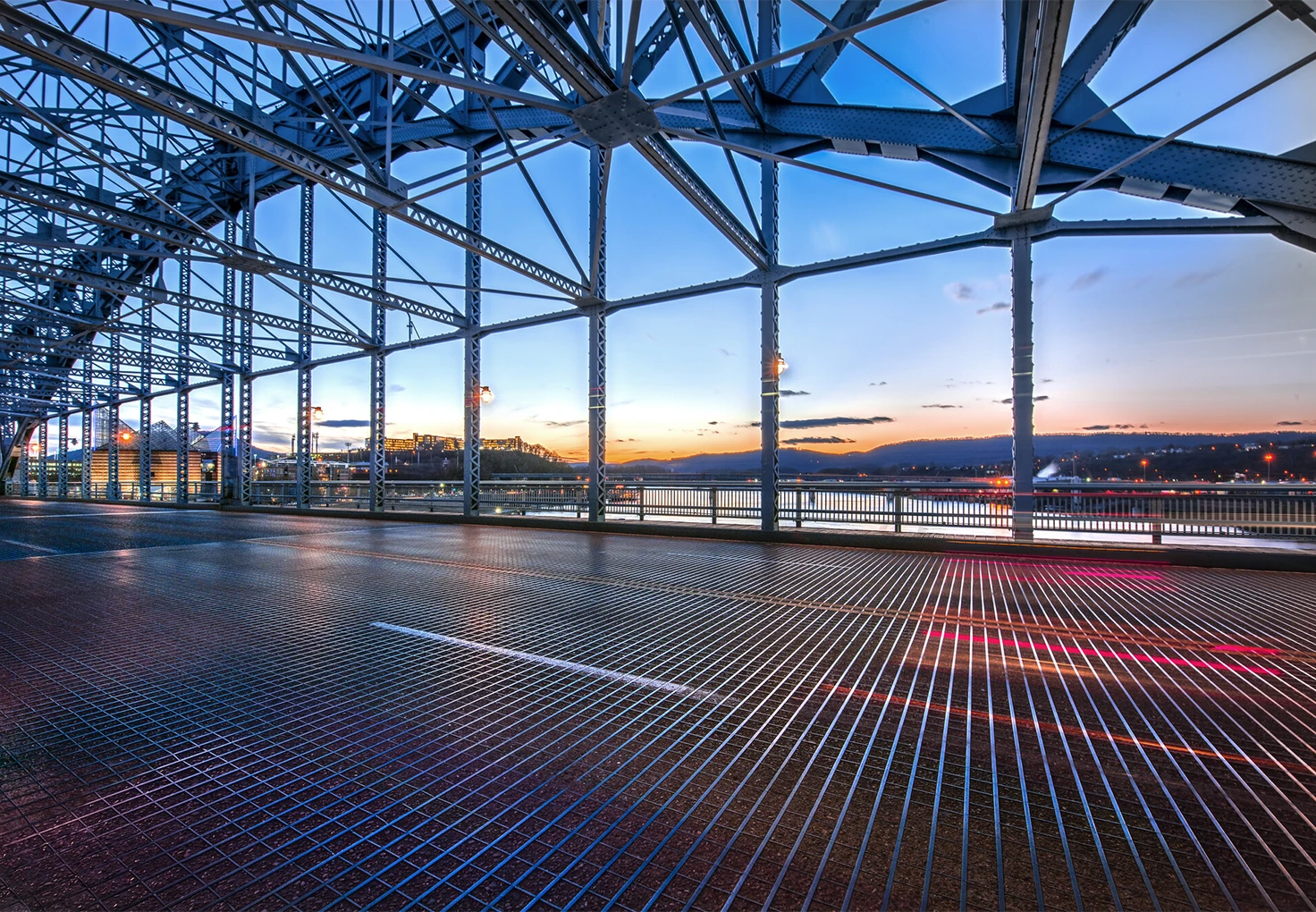 Interior view of a steel bridge at sunset, with visible structural beams and a metal grate floor reflecting colorful light streaks.