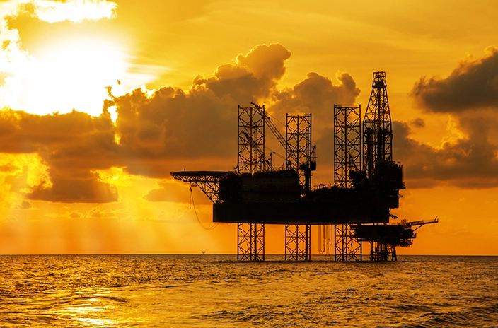 A silhouette of an offshore oil platform against a golden sunset over the ocean.