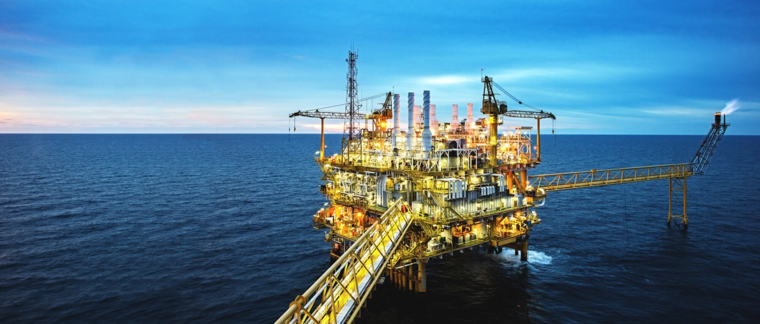 An offshore oil platform brightly illuminated, surrounded by ocean under a blue evening sky.