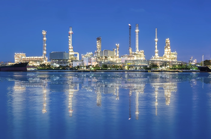A refinery or petrochemical plant at night, with tall structures and chimneys lit up, reflecting on calm water.