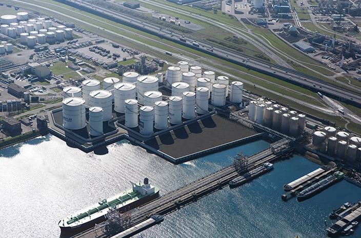 Large storage tanks along a waterfront, connected by pipelines to a ship docked nearby.