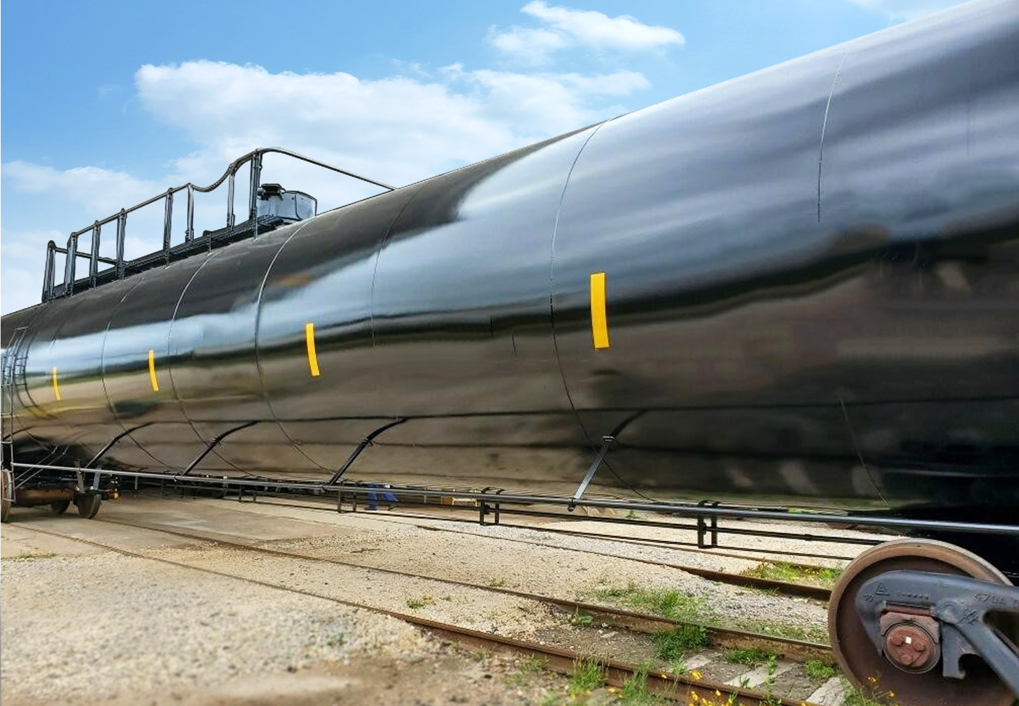 Black rail tank car on tracks, with a shiny surface reflecting the sky. Yellow markings and a ladder on top. Clear day, creating a sleek, industrial feel.