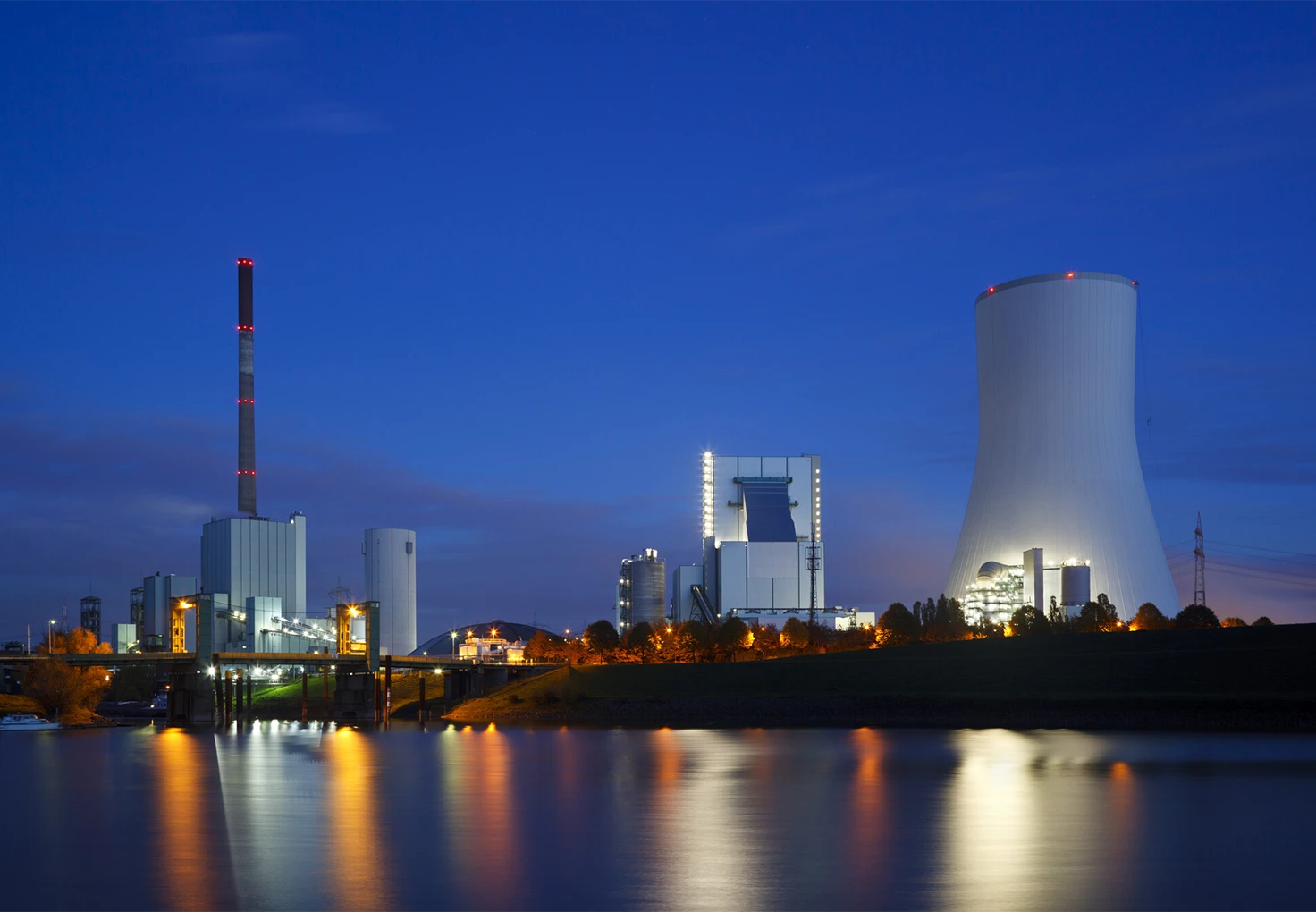 A brightly illuminated power station with a tall cooling tower and chimney, reflecting in the water under a deep blue evening sky.