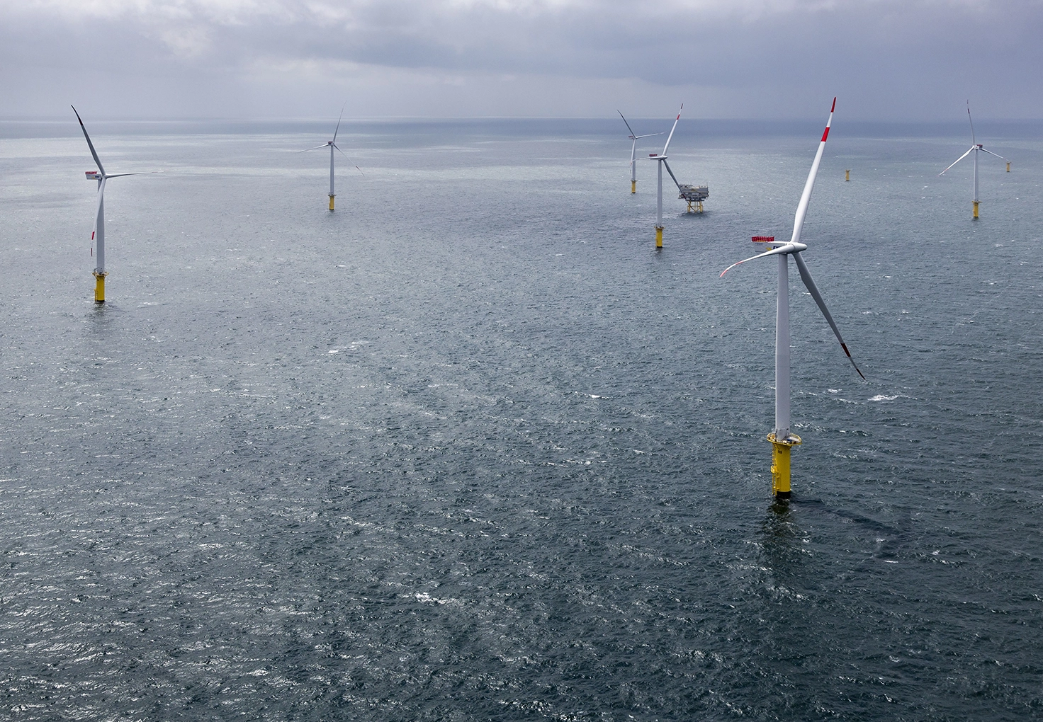 Offshore wind turbines spaced across the ocean under cloudy skies, with a platform visible in the background.
