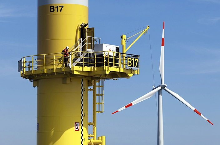 A worker standing on the platform of a yellow offshore wind turbine tower, with the turbine blades visible nearby.