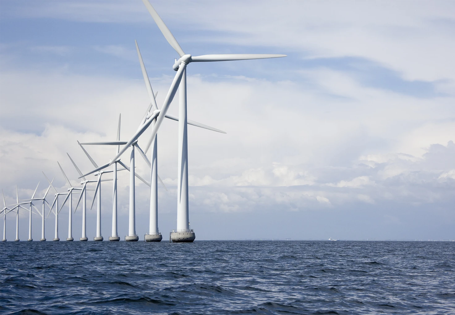 A row of offshore wind turbines generating renewable energy, standing in the ocean under a partly cloudy sky.