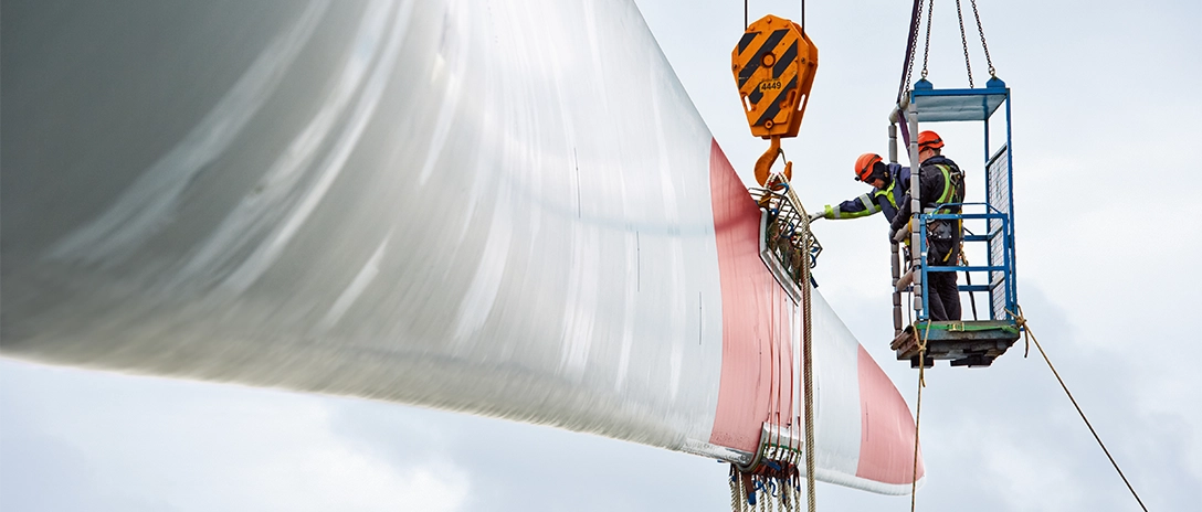 Workers inspecting a wind turbine blade using a crane, with safety harnesses and equipment.