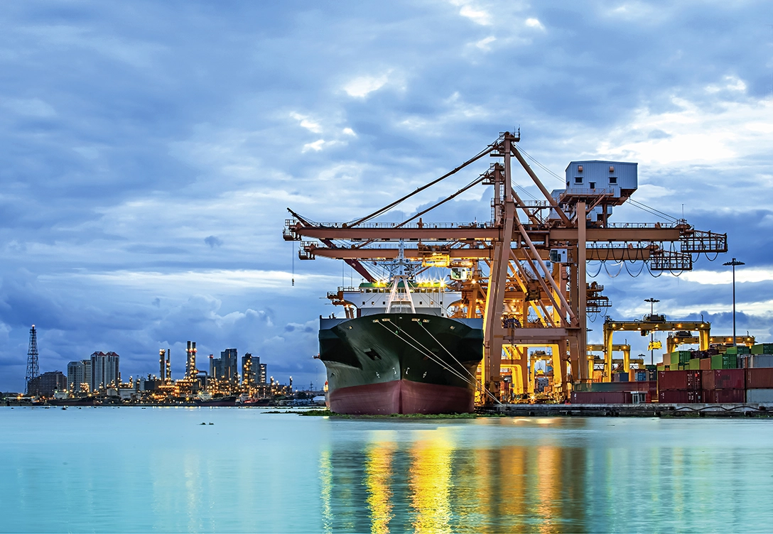 A large cargo ship being loaded at a brightly lit port at dusk, with a city skyline in the background.