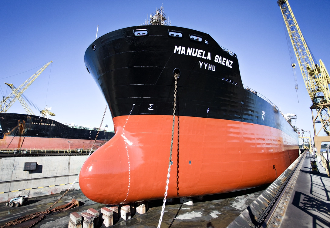 The bow of a large ship named "Manuela Saenz" docked at a shipyard, with cranes and another ship in the background.