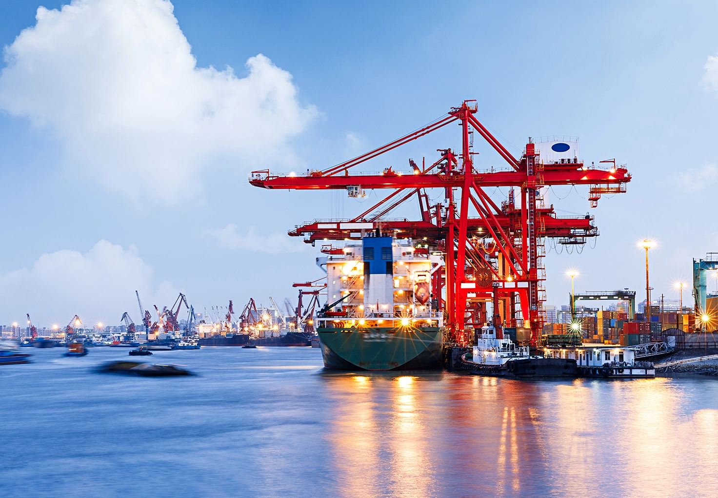 A container ship at a busy dock, surrounded by red cranes, with the dock and ship illuminated, reflecting in the water.