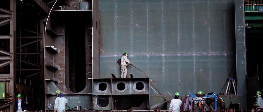 Shipyard workers in hard hats building a large steel hull block.