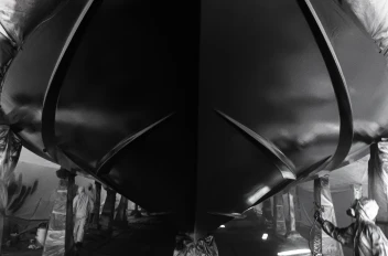 Black-and-white close-up of a yacht’s hull being spray-painted by workers in protective gear.
