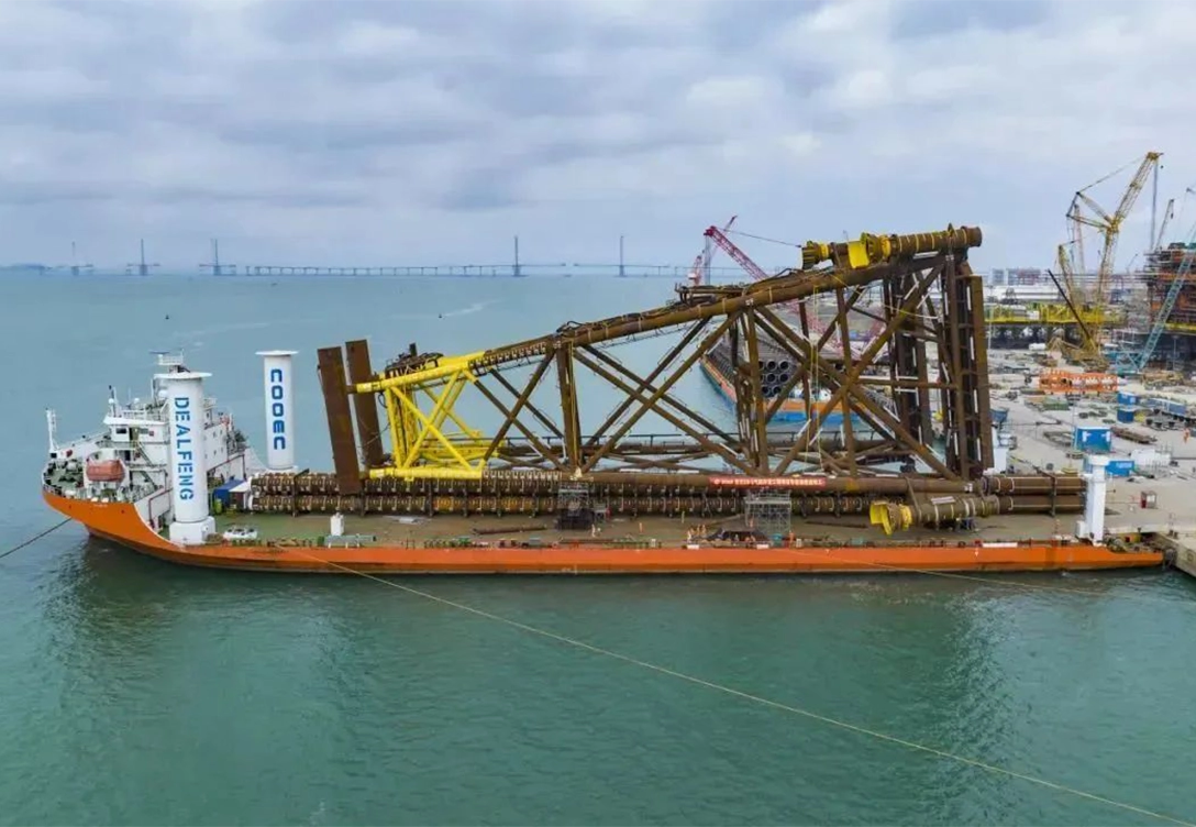 A large barge belonging to COOEC-Fluor Heavy Industries Co., Ltd. carries an offshore platform owned by COOEC, with cranes and industrial facilities visible in the background.