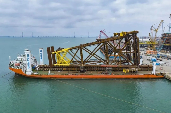 A large barge belonging to COOEC-Fluor Heavy Industries Co., Ltd. carries an offshore platform owned by COOEC, with cranes and industrial facilities visible in the background.