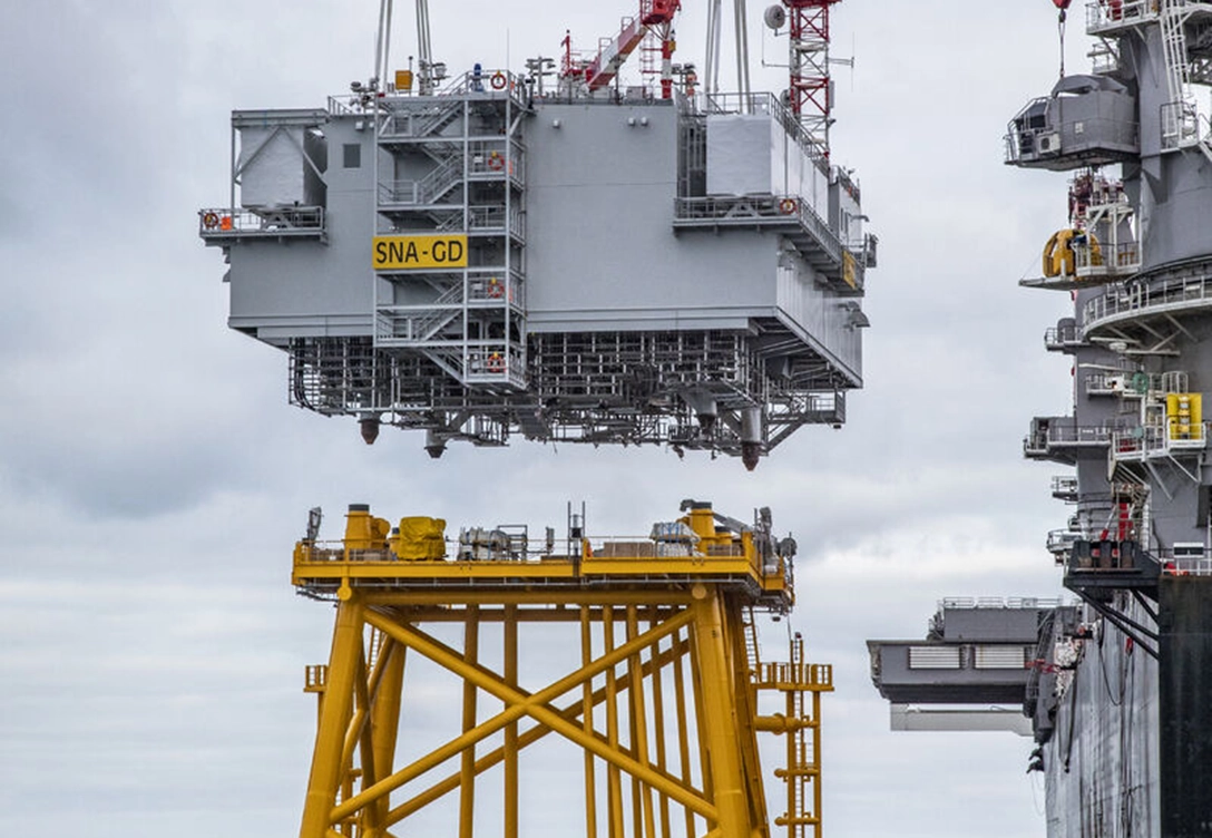 A large offshore substation is being lifted onto a yellow steel foundation at a shipyard.