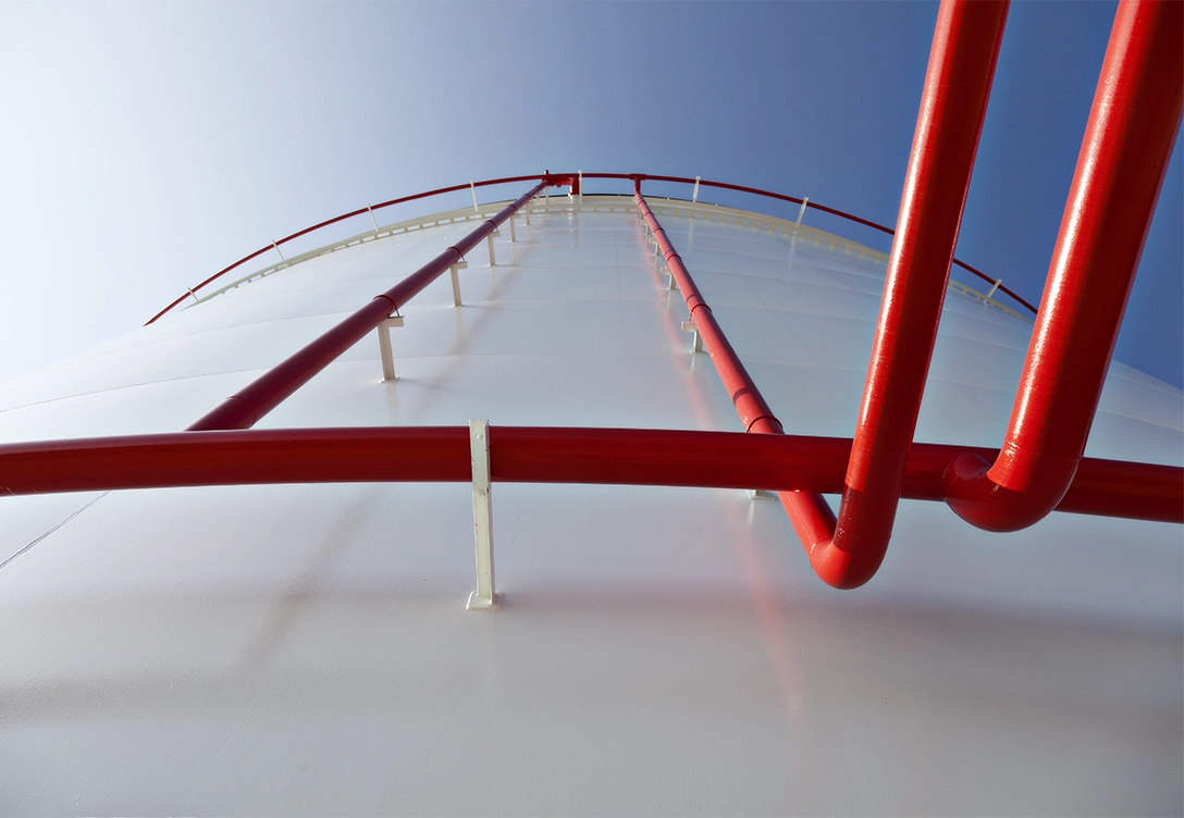 A close-up view looking upward at a large white storage tank with bright red pipes extending vertically against a blue sky.