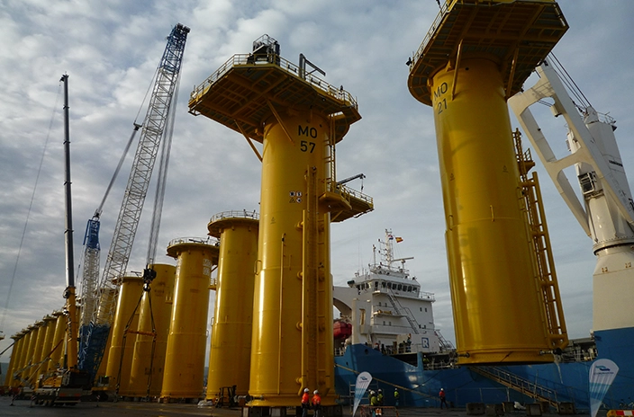 Yellow-painted foundation structures for the Merkur Windfarm lined up on a dockside.
