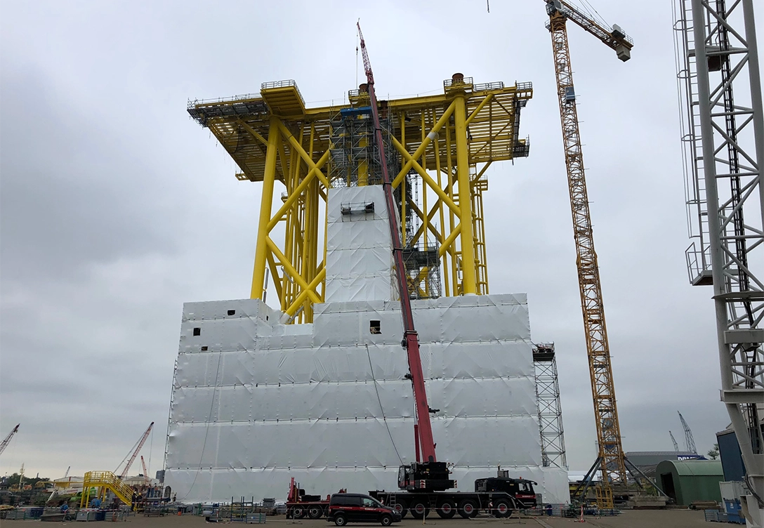 Offshore wind station foundation under construction with scaffolding and tarps, cranes are actively being used.